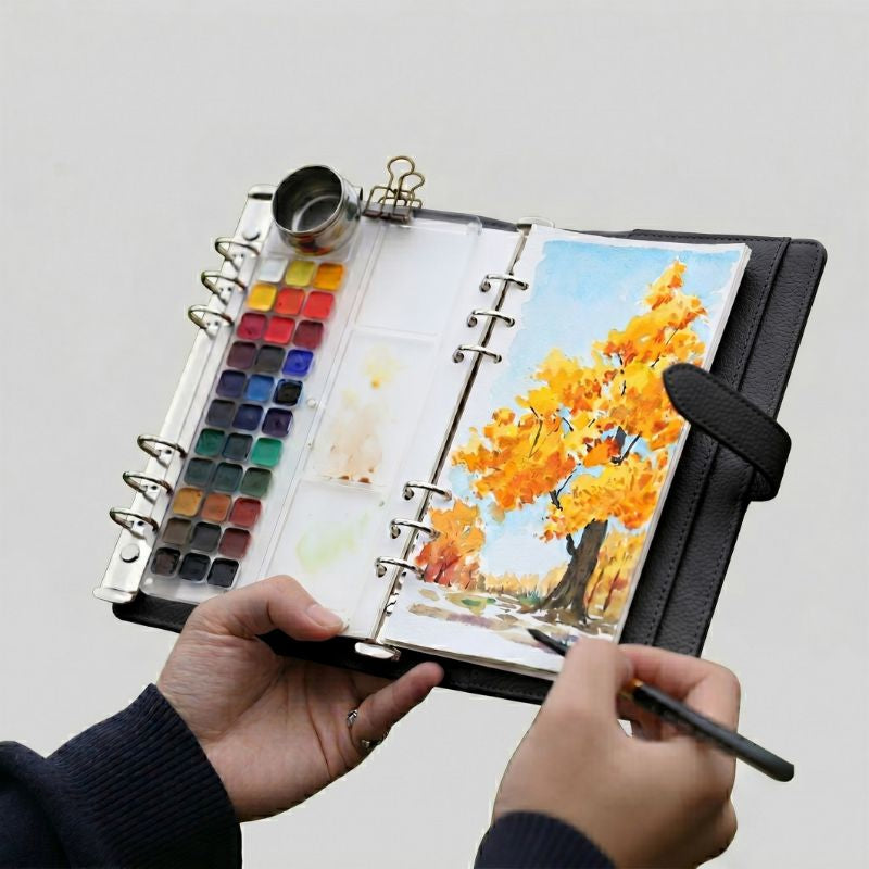 Hands holding the open Classic Black Voyager watercolor kit, painting an autumn tree with the 33-pan palette and water pot