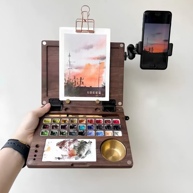 Open walnut Master Edition station held in hand with a sunset painting clipped to the lid, a 27-pan palette and brass water pot, and a phone mounted on the adjustable arm as a reference